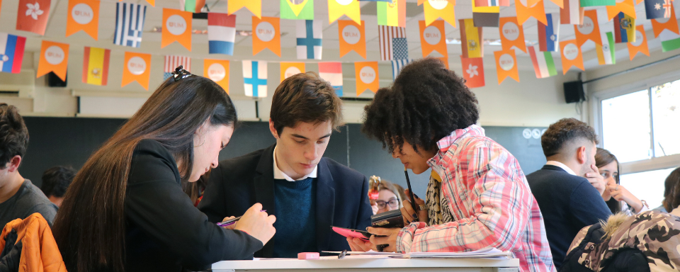 Tres alumnos dialogan alrededor de una mesa. Arriba se ven banderines con bandereas de distintos países.