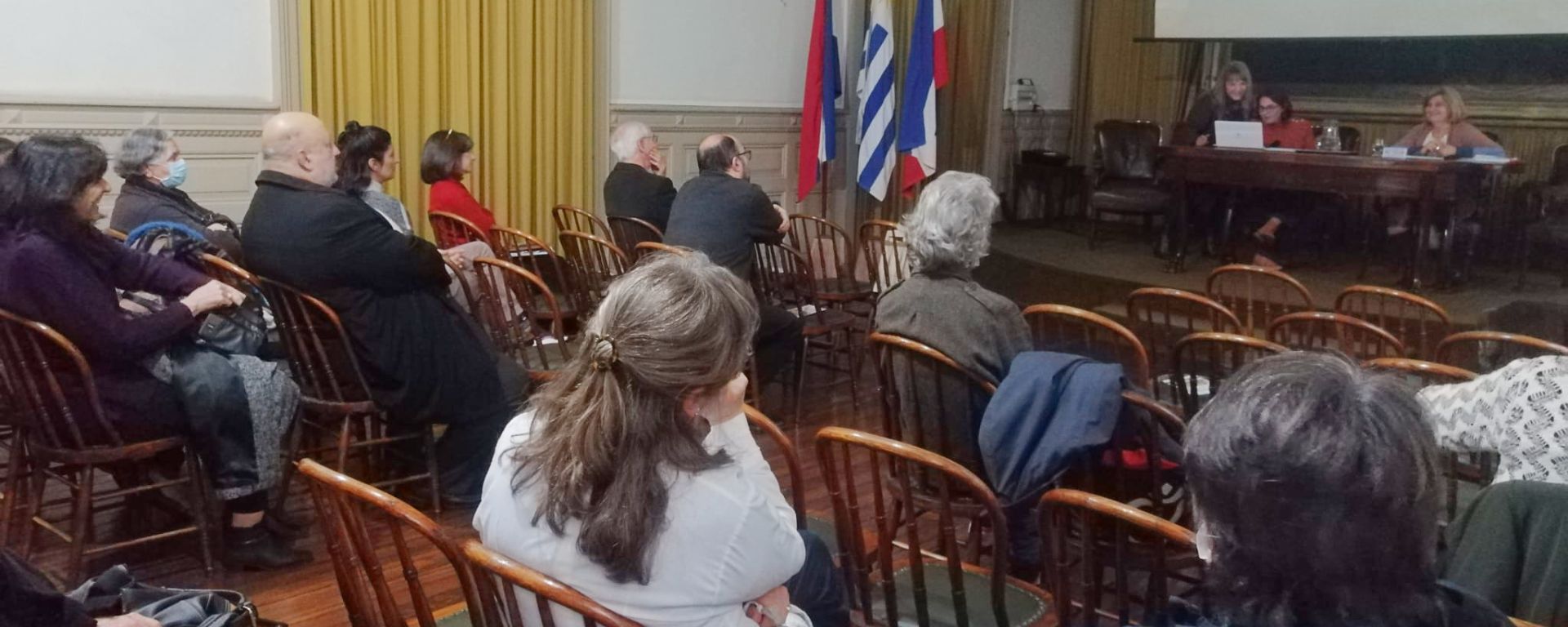 Imagen de conferencia en salón con público de espaldas y conferencistas en mesa frontal sobre escenario