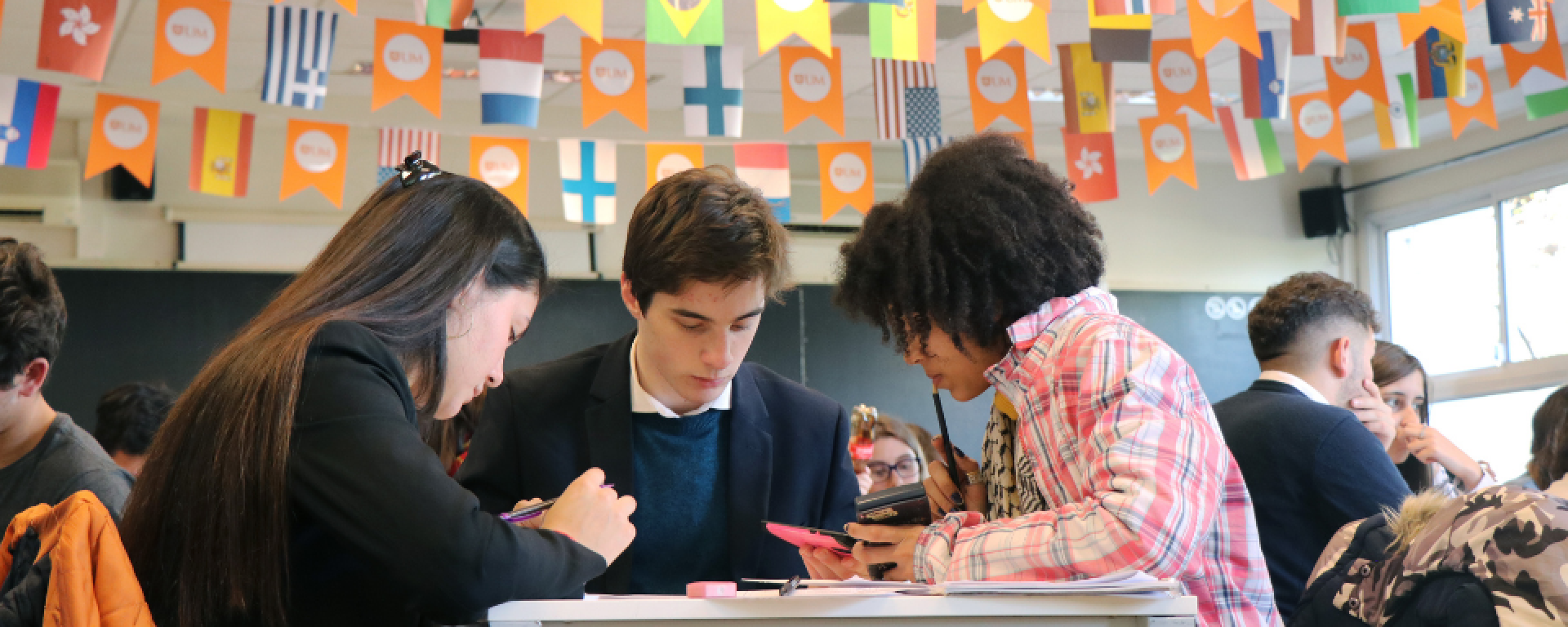 Tres alumnos dialogan alrededor de una mesa. Arriba se ven banderines con bandereas de distintos países.